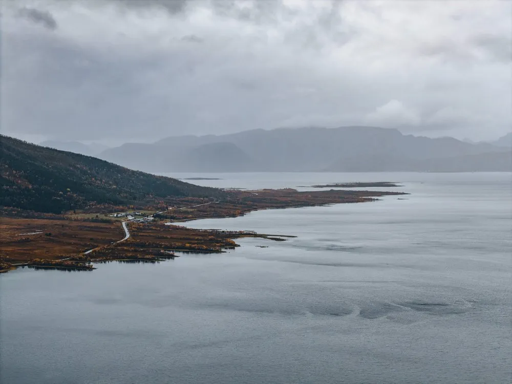 Eine Landzunge mit einer Strasse zieht sich ins graue Wasser, dahinter Berge im Regen, aus der Luft unterwegs nach Andøya