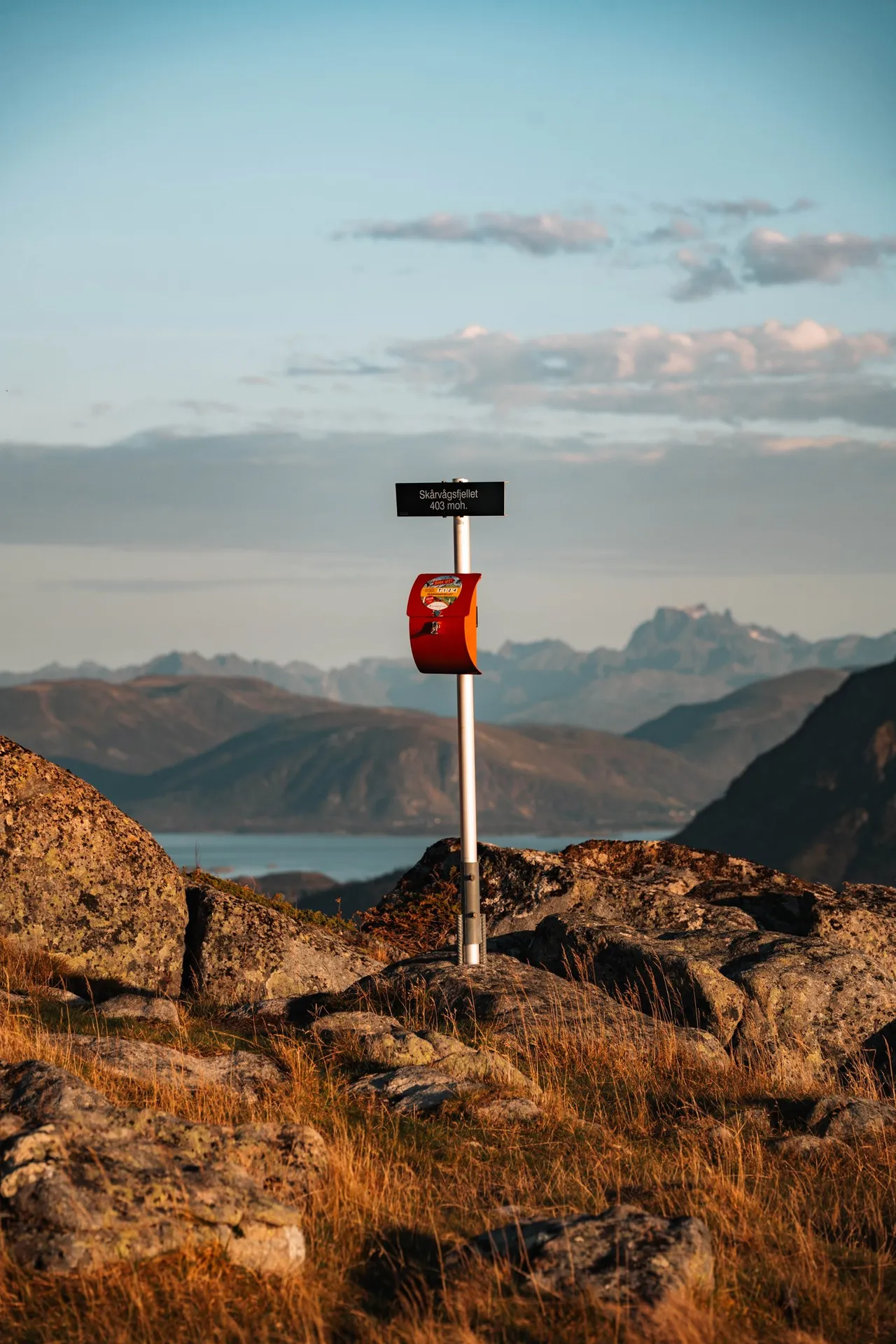 Das Gipfelschild «Skårvågsfjellet 403 moh.» mit dem roten Kasten von «Stikk UT!» zwischen Felsblöcken, dahinter Berge und Wasser