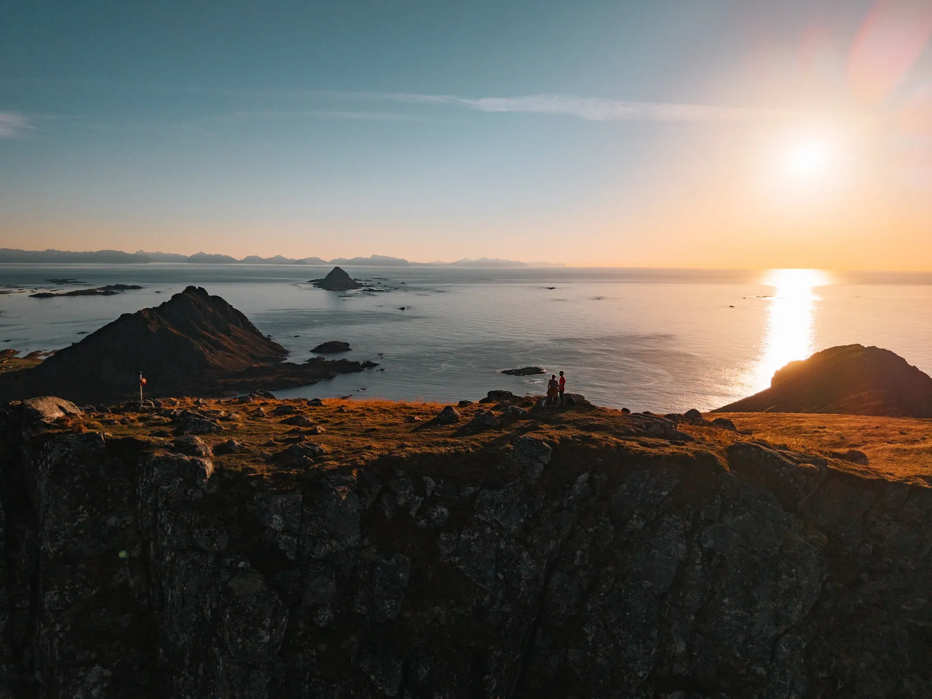 Wir als kleine Figuren am Rand des Skårvågsfjellet, links der spitze Gullstabben, in der Mitte die Insel Gaukværøya und am Horizont die Lofoten, rechts die tief stehende Sonne