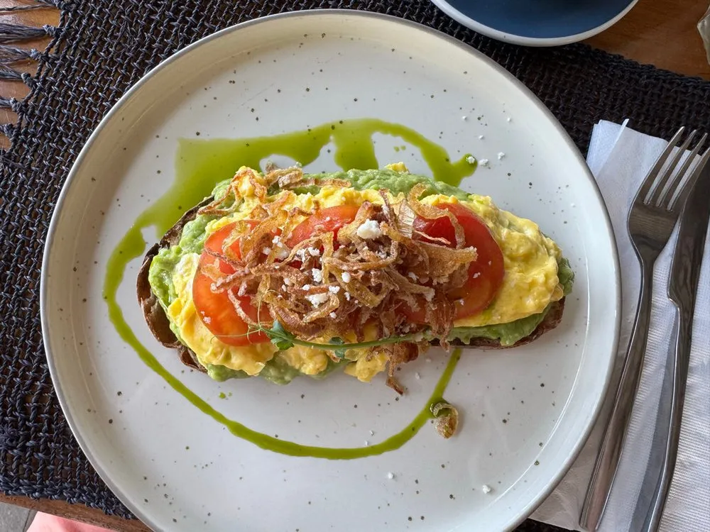 Toast mit Avocado, Rührei, Tomatenscheiben und knusprigen Zwiebelstreifen auf einem hellen Teller mit dunklen Sprenkeln