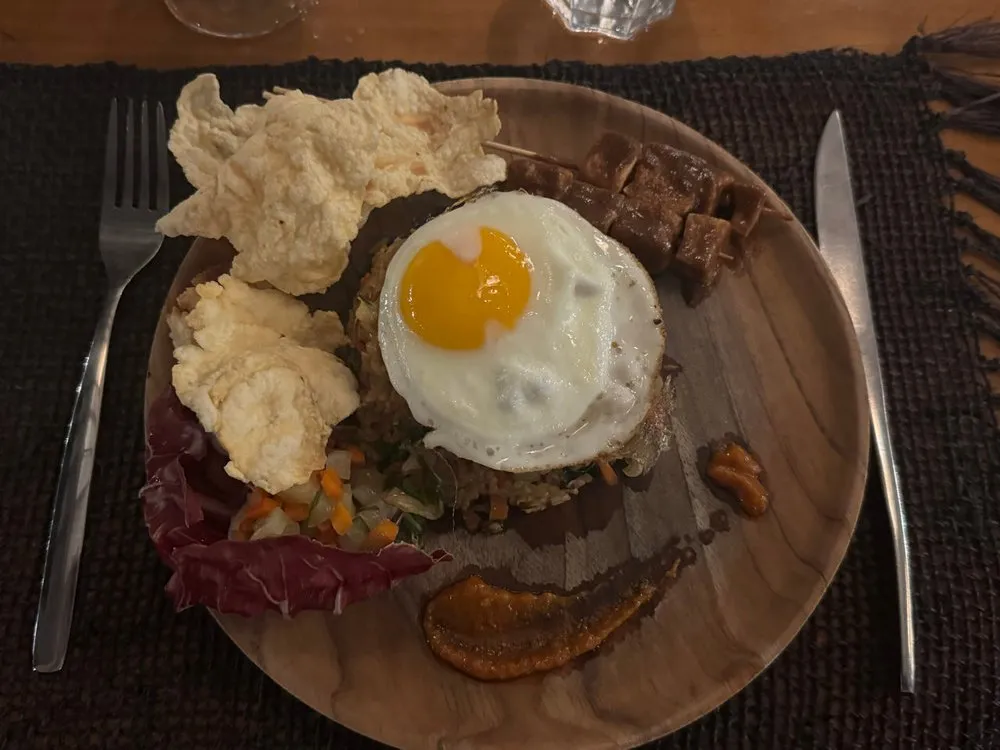 Nasi Goreng with a fried egg on a wooden plate, next to it two light crackers, diced vegetables and a small skewer