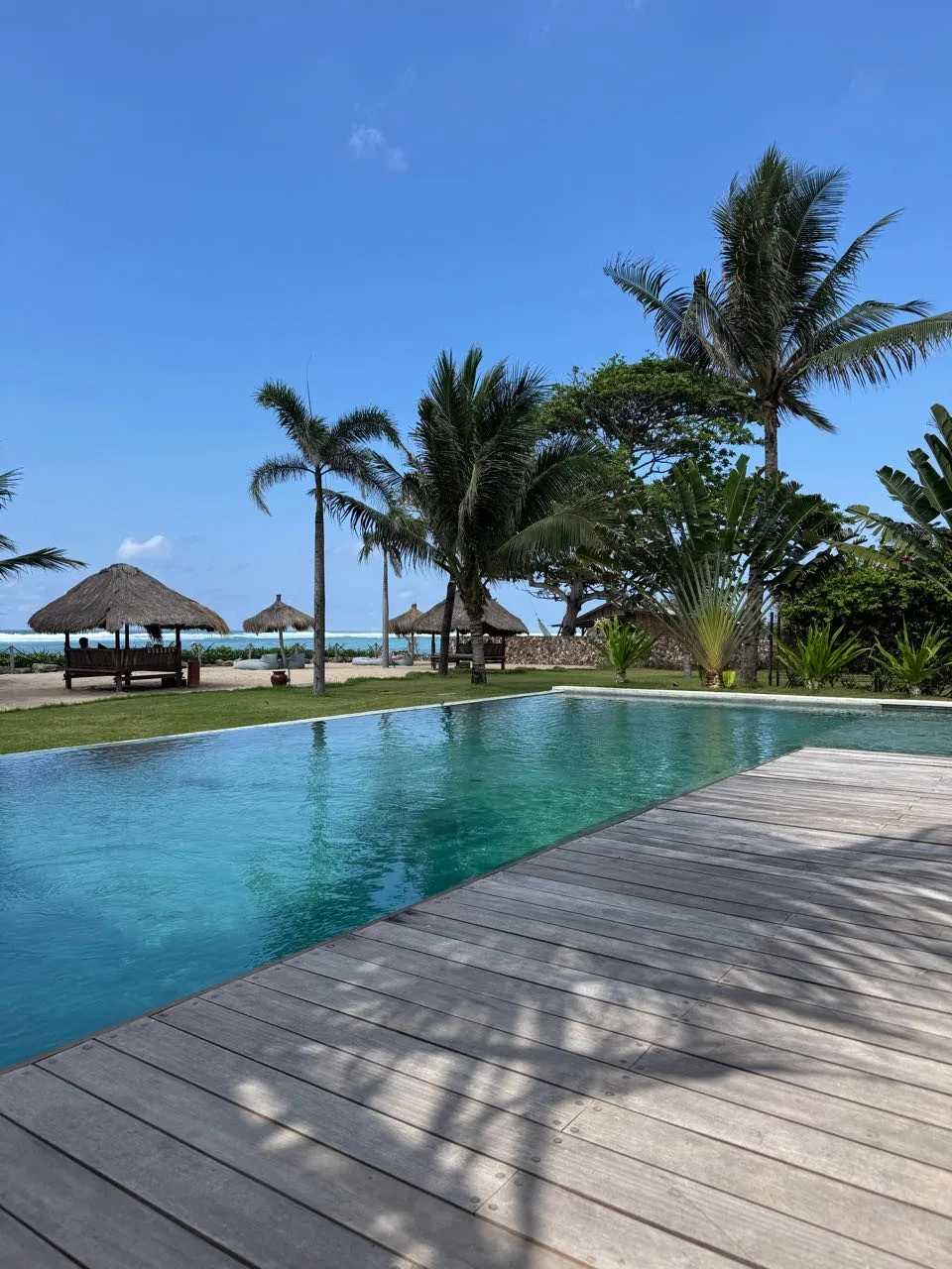 Ein Pool mit Holzdeck unter blauem Himmel, dahinter Palmen, Pavillons mit Strohdach und in der Ferne das Meer