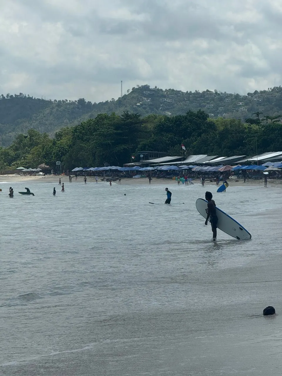 Viel Betrieb am Strand von Selong Belanak, blaue Schirme am Ufer, im flachen Wasser Surfer, einer trägt sein Brett