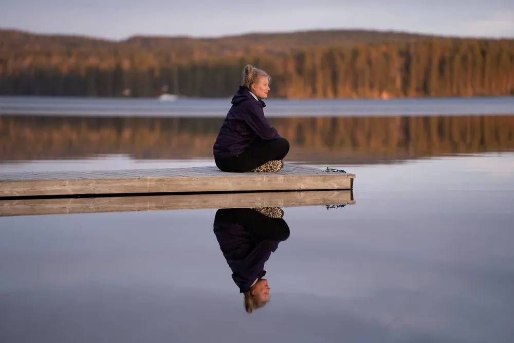 Eine Frau in dunkler Jacke sitzt am Stegrand, ihr Spiegelbild im Wasser, dahinter Wald im warmen Licht