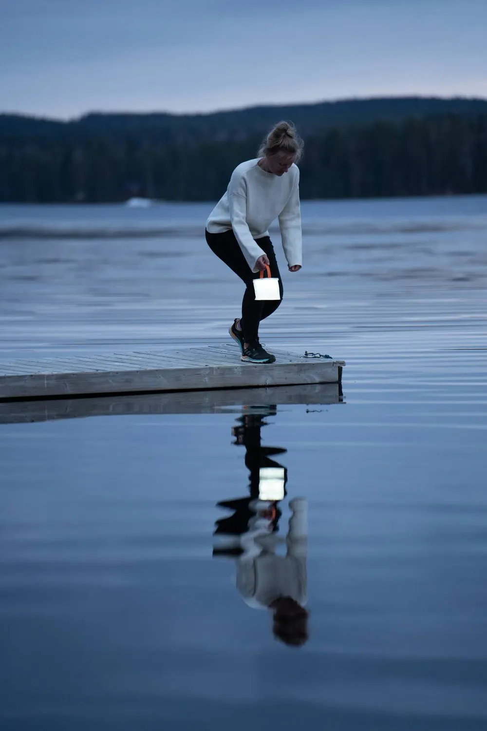 Eine Frau in weissem Pullover beugt sich auf dem Steg mit einer leuchtenden Lampe in der Hand vor, ihr Spiegelbild im ruhigen Wasser