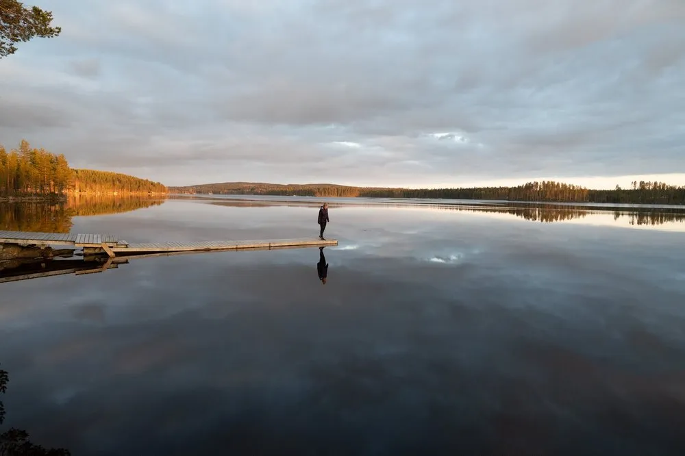 Eine Person steht am Ende eines langen Stegs, Wolken und Ufer spiegeln sich im Wasser