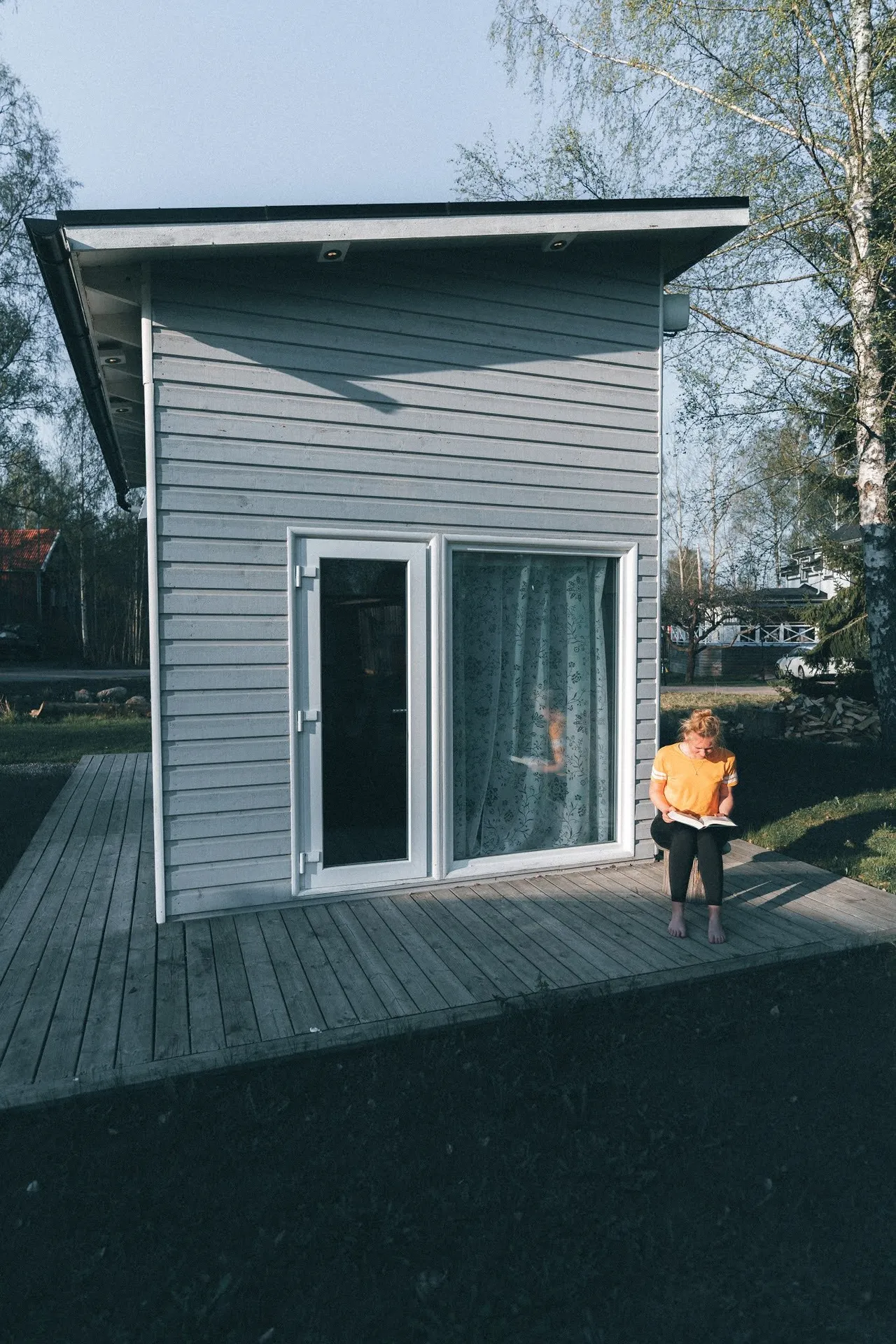 The tiny house with grey wood cladding and a terrace, a person in an orange top sitting in front of it reading