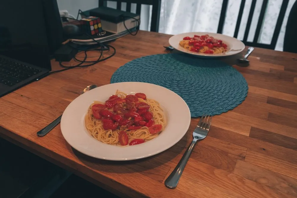 Zwei Teller Spaghetti mit Cherrytomatensauce auf einem Holztisch, daneben ein Laptop und ein Zauberwürfel