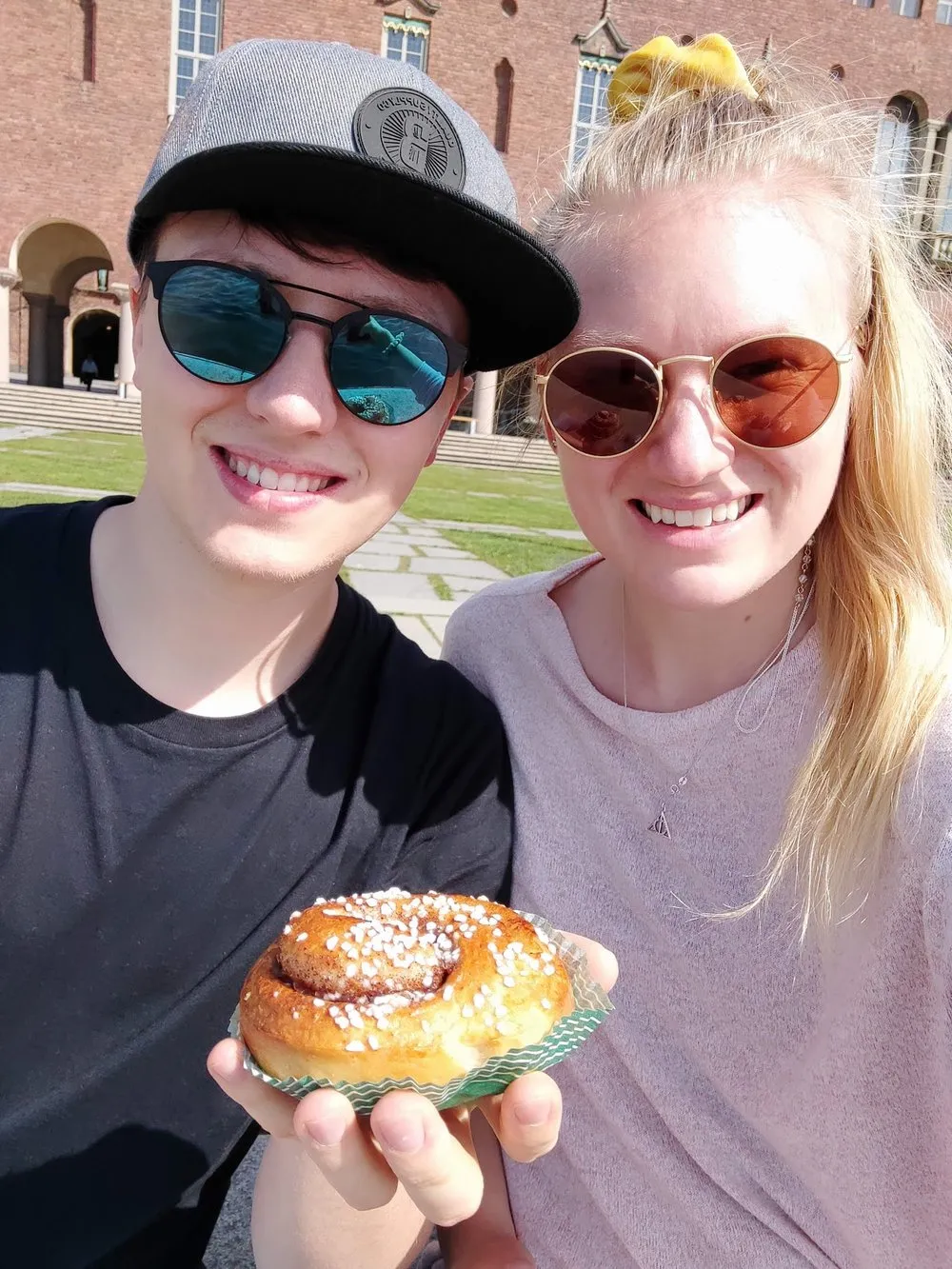 Zwei Personen mit Sonnenbrillen halten zum Dessert am Stadhus eine Zimtschnecke in die Kamera, dahinter eine Backsteinfassade mit Rundbögen