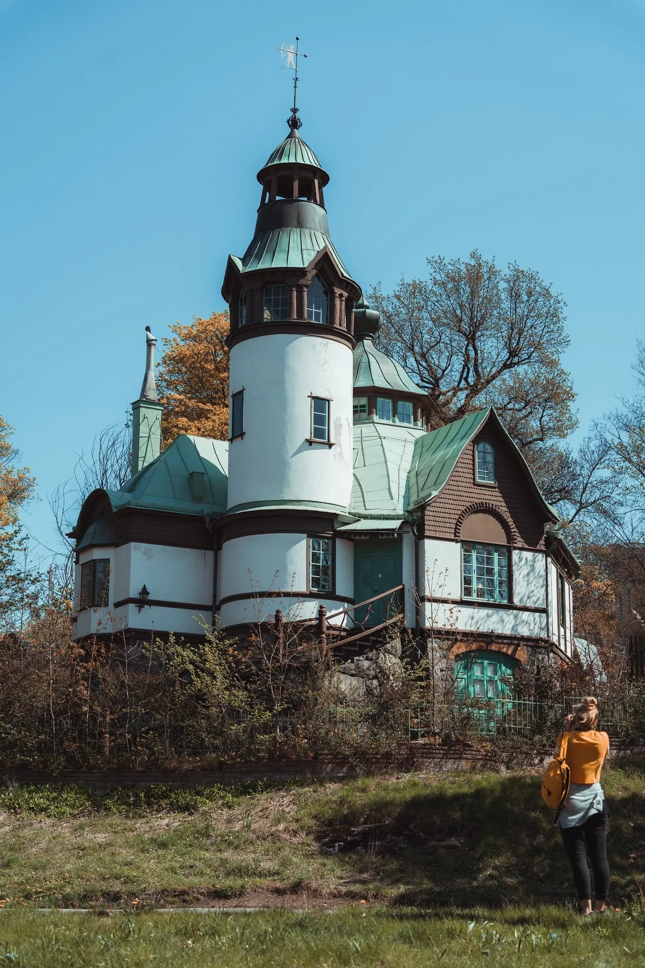 Villa with a white round tower, green copper roof and wood cladding, in front of it a person in an orange top taking photos in the grass