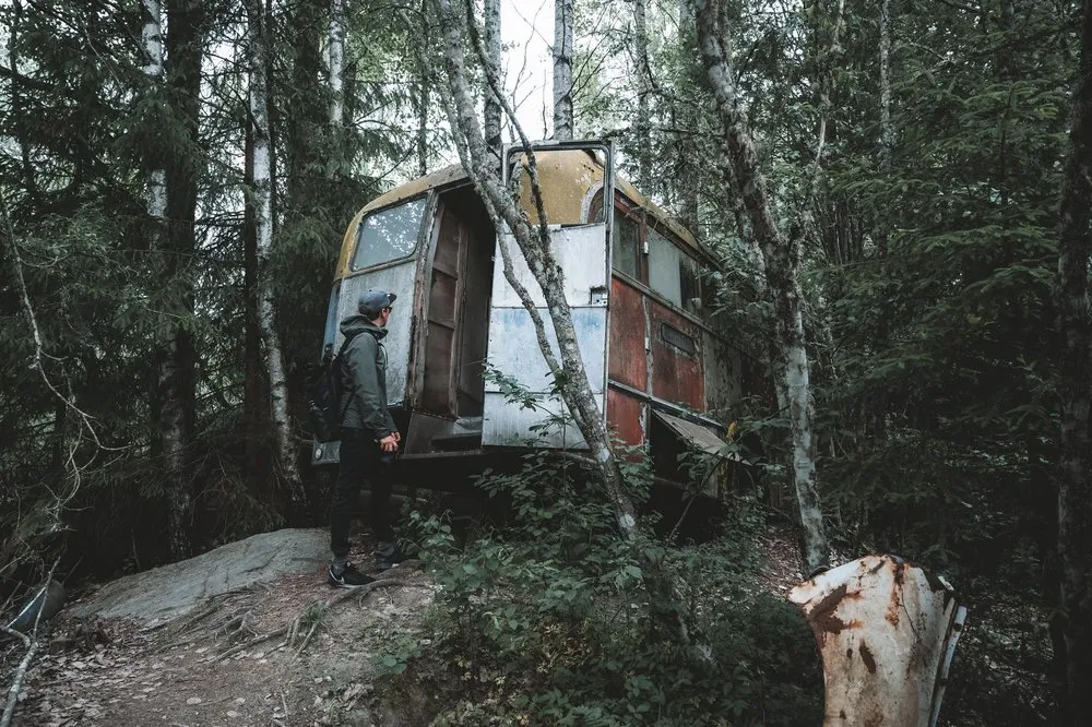 Ein Mann mit Kappe und Rucksack steht auf einem Felsen vor einem verrosteten Wohnwagen im Wald