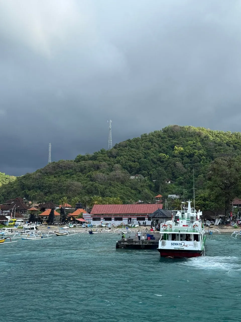Ein Hafen mit Auslegerbooten, einem weissen Boot und roten Dächern vor einem grünen Hügel, darüber dunkle Wolken