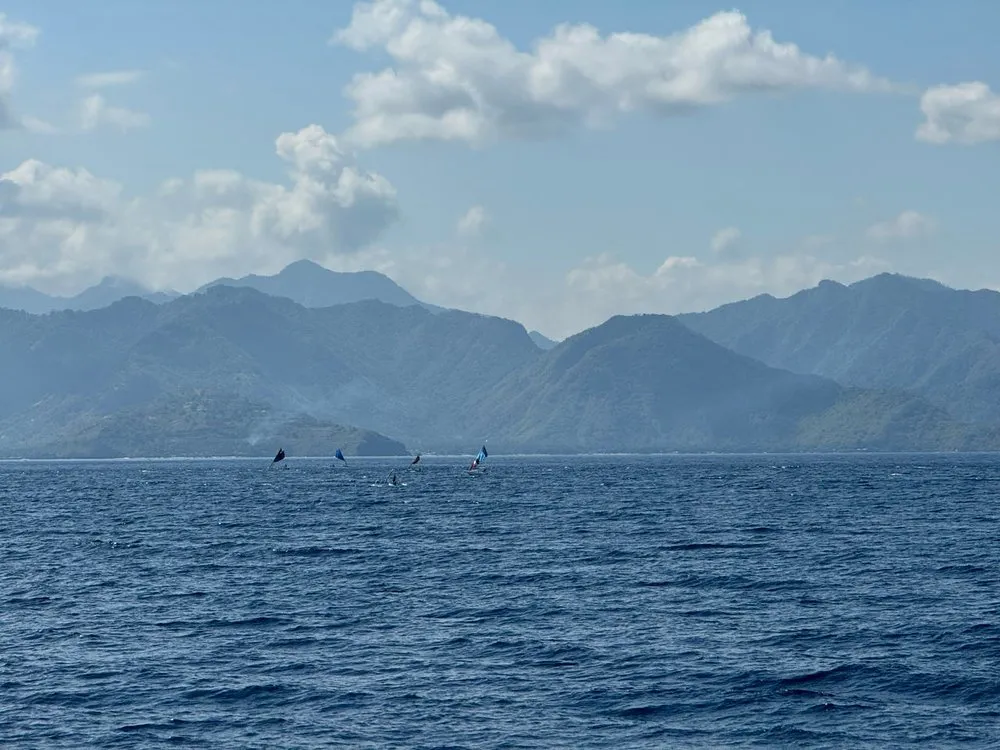 Blaue Bergketten über dem Meer, davor einige kleine Boote mit dunklen Segeln