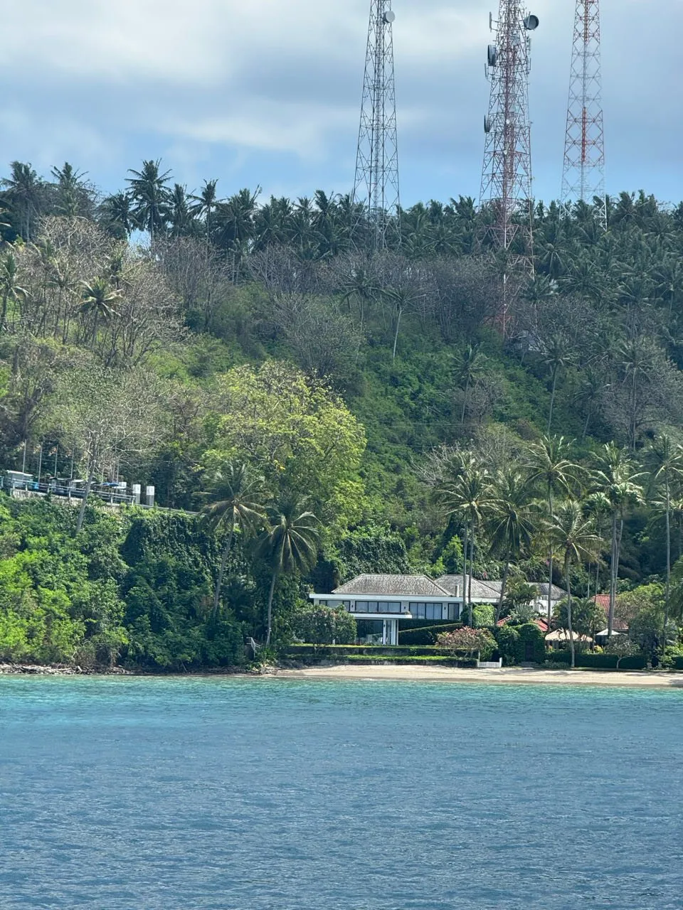 Bewaldeter Hang mit Palmen und Funkmasten, am Ufer ein Haus mit grauem Dach vor einem schmalen Sandstrand