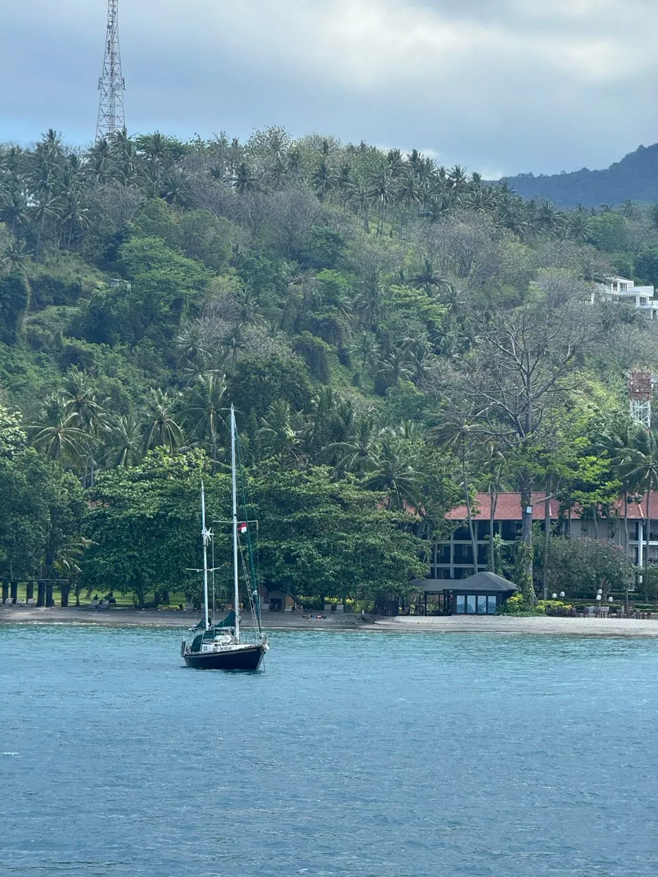 Ein Segelboot liegt vor Anker vor einer palmenbewachsenen Küste mit einem Gebäude mit roten Dächern