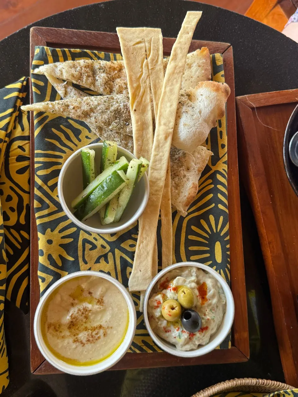 Holzbrett mit Fladenbrot, Gurkenstreifen und zwei Dips, einer mit Olivenstücken, auf einem gelb-schwarz gemusterten Tuch