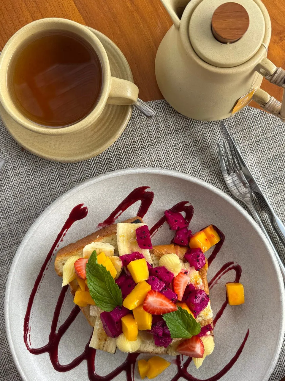 Brotscheibe mit Mango, Banane, Erdbeeren und pinken Fruchtstücken auf dunkelroter Sauce, daneben eine Tasse Tee und eine Kanne