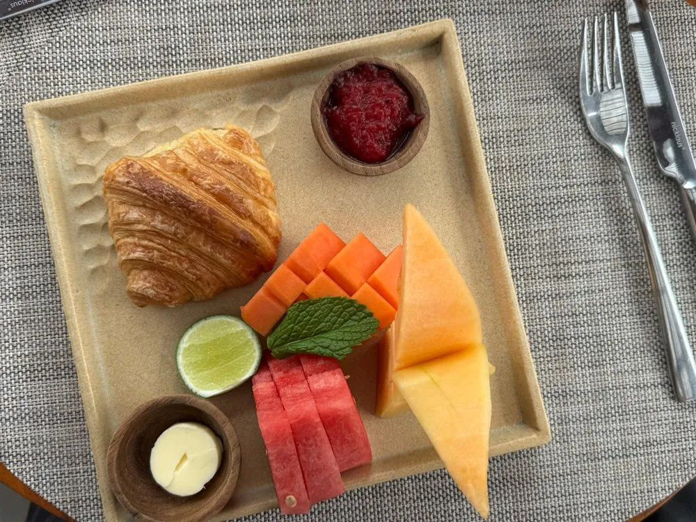 Croissant mit Butter und roter Konfitüre neben Wassermelone, Melone, orangen Fruchtstücken und einer halben Limette