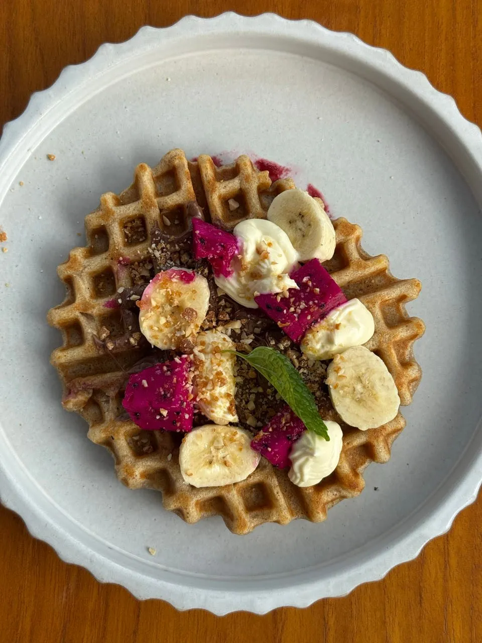 Waffel mit Bananenscheiben, pinken Fruchtstücken, Cremetupfern, gehackten Nüssen und Minze auf einem hellen Teller