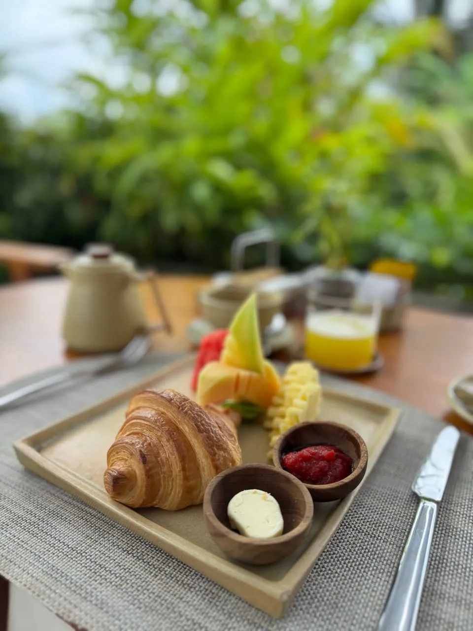 Croissant mit Butter und roter Konfitüre in Holzschälchen, dahinter Fruchtscheiben, ein Glas Saft und eine kleine Kanne vor grünem Laub