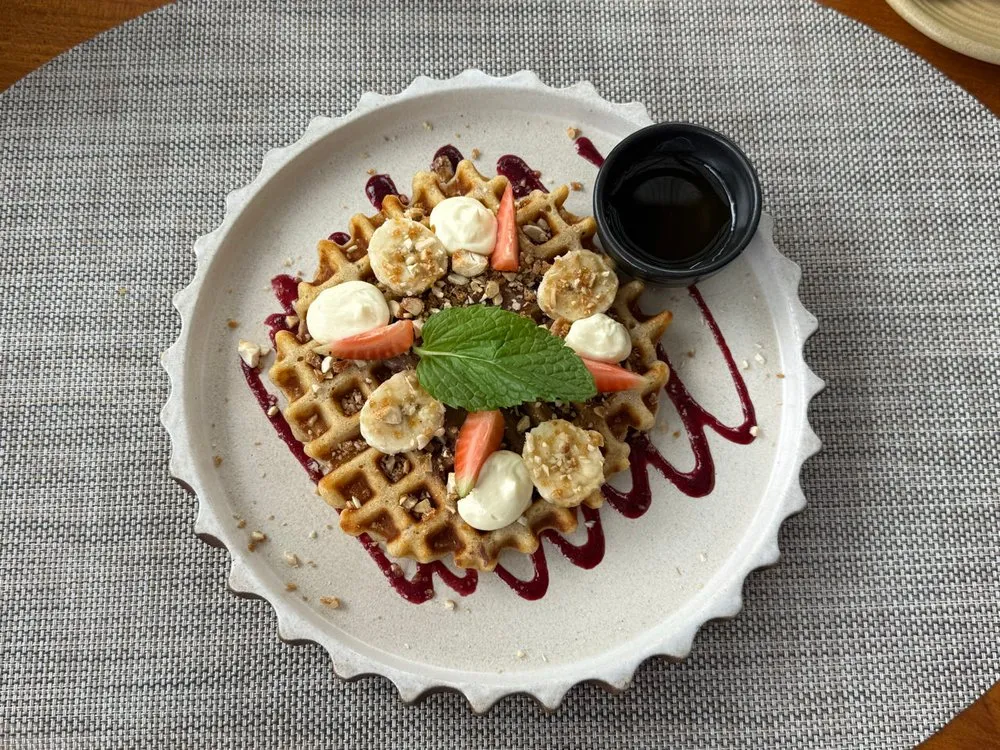 Waffel mit Bananenscheiben, Cremetupfern, gehackten Nüssen und einem Minzblatt, daneben ein Schälchen mit dunkler Sauce