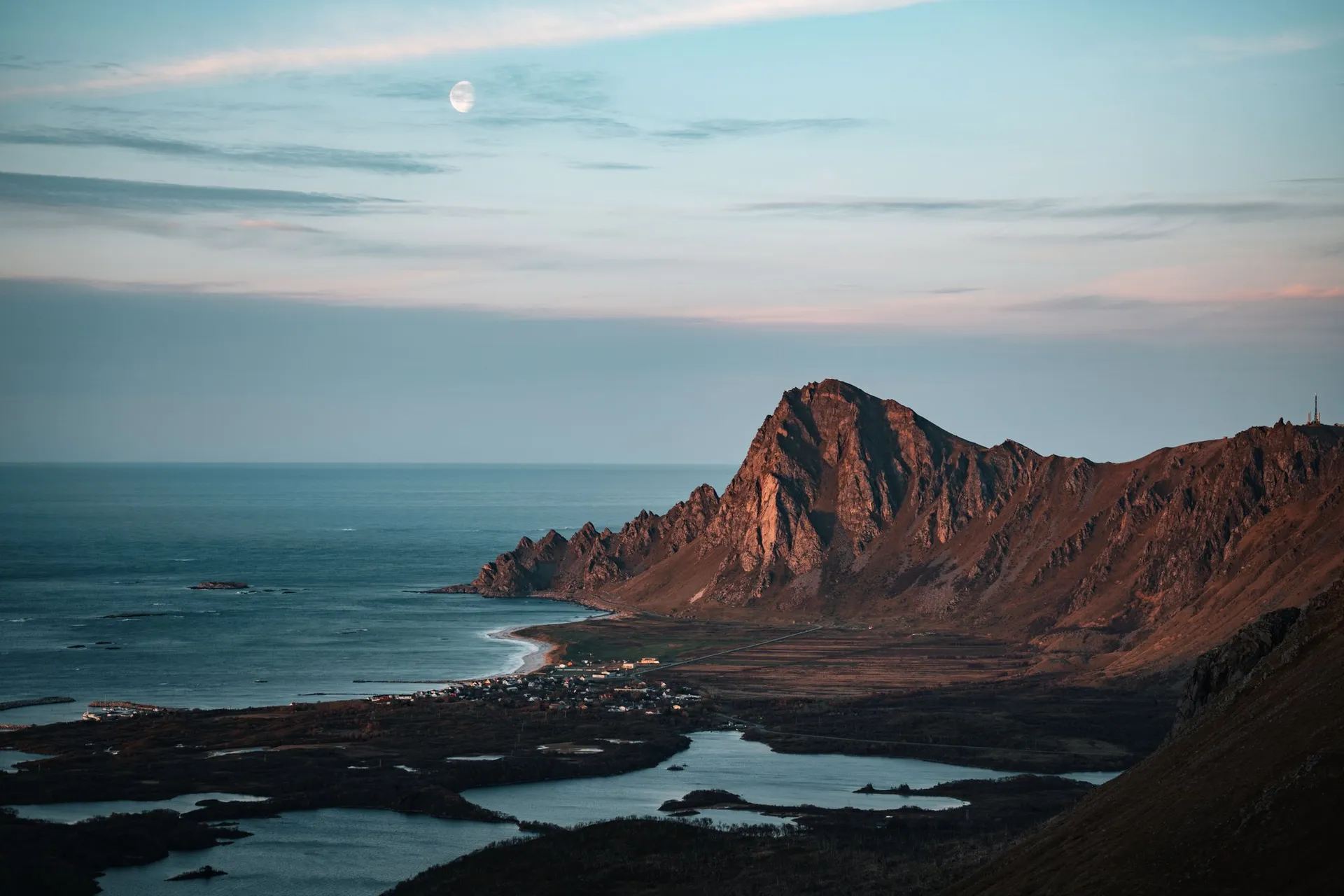 The moon above a mountain in the evening light, a village, lakes and the sea below, on the way down Måtind