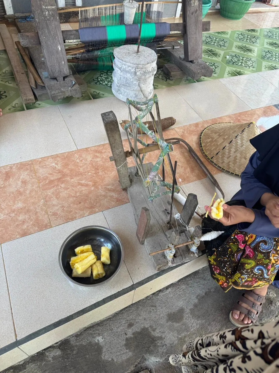 A wooden spinning wheel, behind it a loom with blue and green striped threads, on the left a metal bowl with yellow pieces