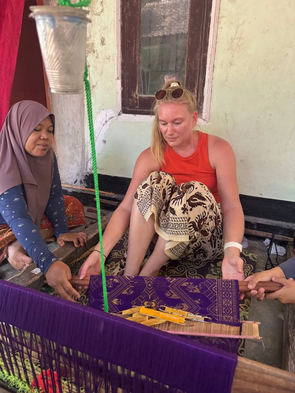 Annina sits with a woman in a headscarf in front of a loom with violet patterned fabric
