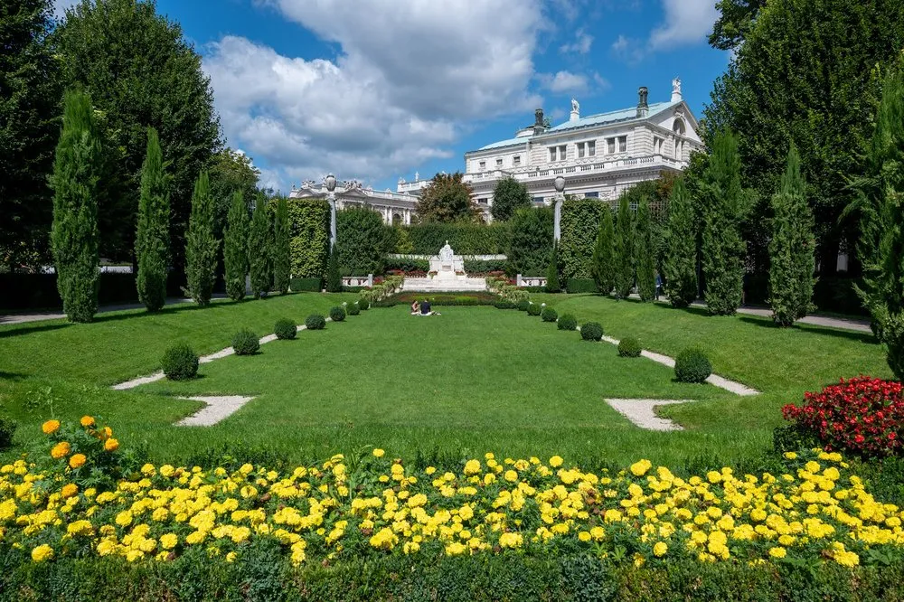 Gepflegte Parkanlage mit Rasen und gelb blühendem Beet, in der Mitte ein weisses Denkmal, dahinter ein Gebäude mit grünem Dach