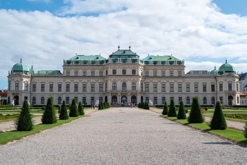 Die Vorderseite von Schloss Belvedere mit grünen Dächern, davor ein Kiesweg und Zierbäume unter blauem Himmel mit Wolken