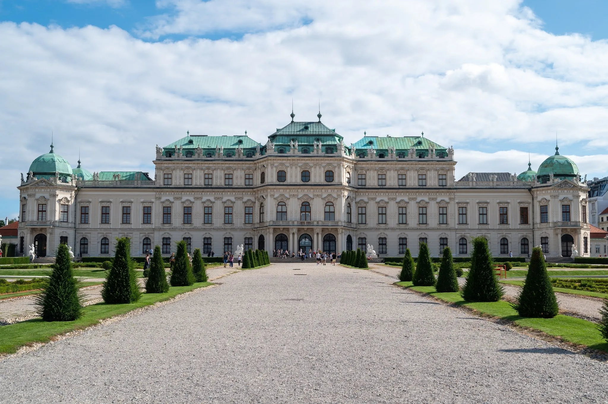 Die Vorderseite von Schloss Belvedere mit grünen Dächern, davor ein Kiesweg und Zierbäume unter blauem Himmel mit Wolken