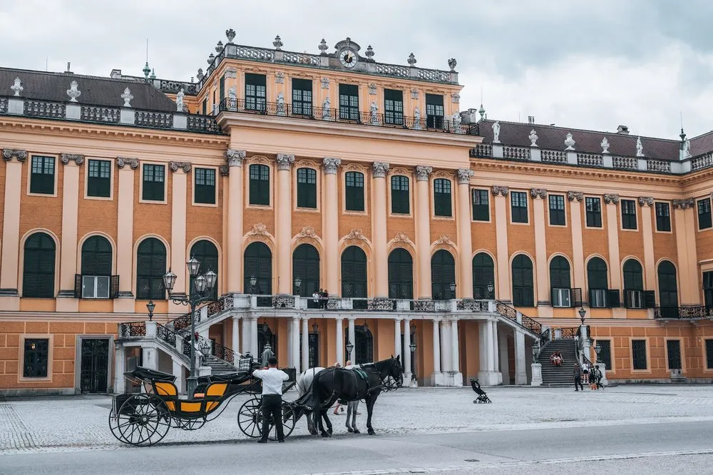 Die orange Fassade von Schloss Schönbrunn mit Treppe und Arkaden, davor eine Pferdekutsche mit einem Mann daneben