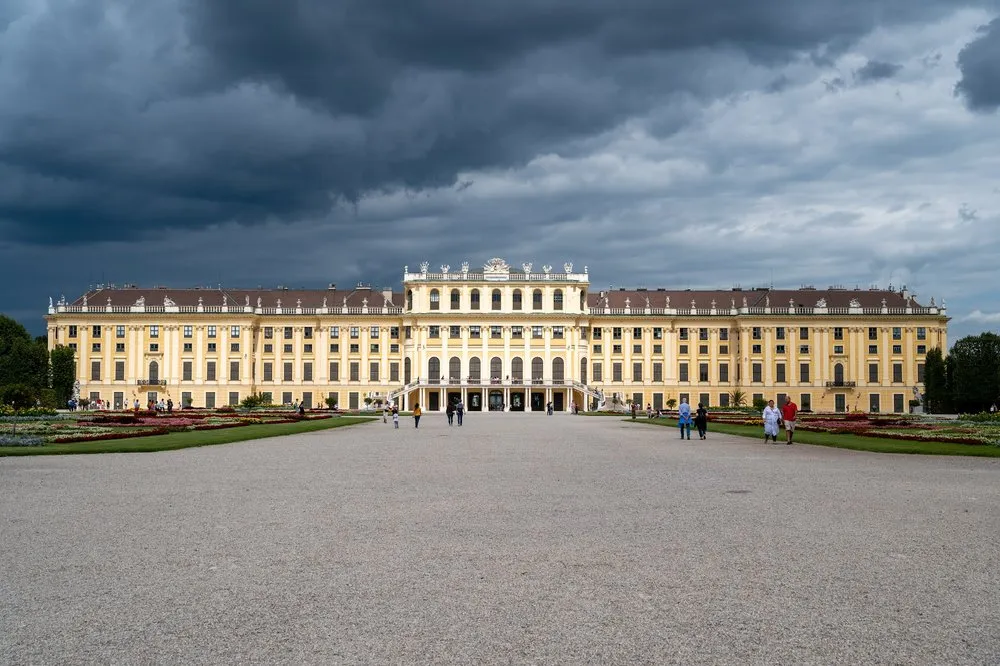 Schloss Schönbrunn unter dunklen Gewitterwolken, davor der weite Kiesplatz mit einigen Besuchern