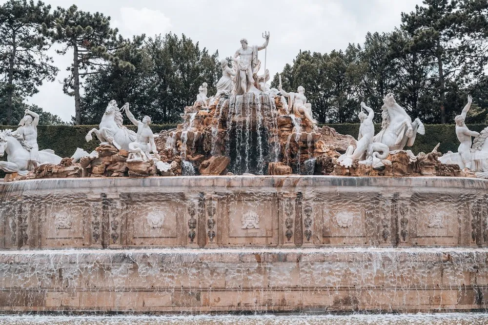 Ein Brunnen mit weissen Figuren, Pferden und einer Figur mit Dreizack auf einem Felsaufbau, das Wasser fällt über die Stufen