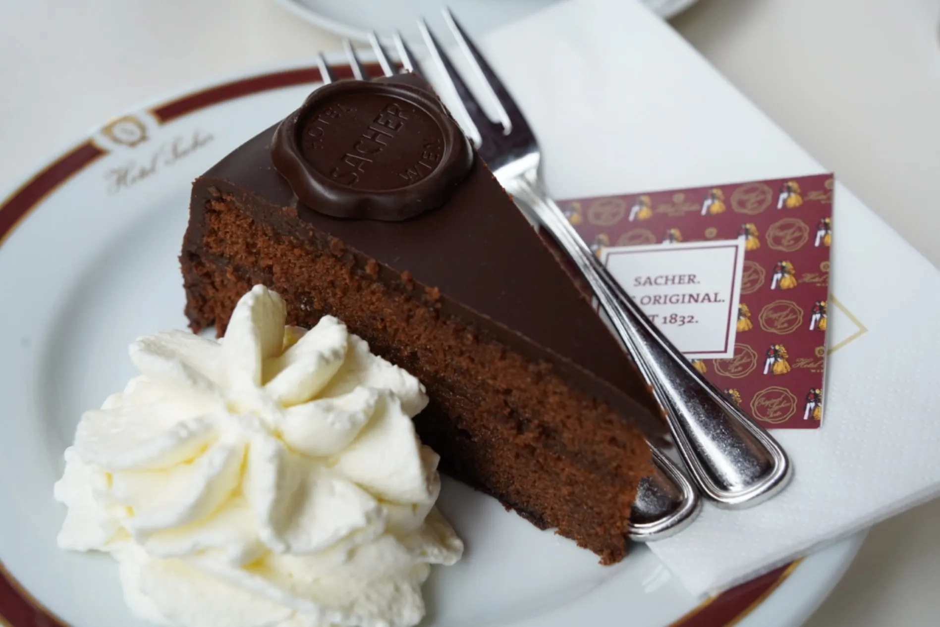 Ein Stück Sachertorte mit Schokoladenglasur und rundem Sacher-Siegel, daneben Schlagsahne und zwei Gabeln auf einem Teller
